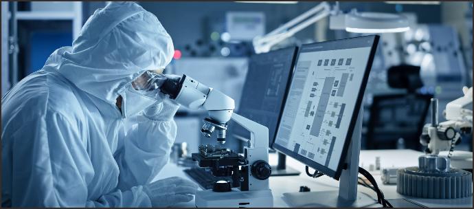 Researcher in protective gear working with a microscope and monitoring data on a computer screen, illustrating the product custom research and development process from technical needs assessment to performance testing and improvement